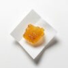 An extreme macro close-up of a small portion of Cannatrek T50 Topaz Bubble Hash concentrate resting on a clean white pharmaceutical weigh paper, soft diffused overhead studio lighting, sterile white laboratory-style background, camera positioned directly overhead at a very slight angle, ultra-high detail rendering capturing the golden-amber colour, surface texture, and trichome-rich consistency of the hash material, clean scientific product documentation photography mood with no lifestyle elements