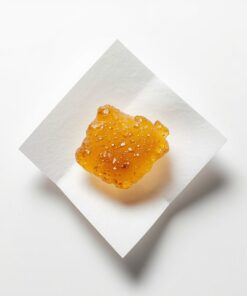 An extreme macro close-up of a small portion of Cannatrek T50 Topaz Bubble Hash concentrate resting on a clean white pharmaceutical weigh paper, soft diffused overhead studio lighting, sterile white laboratory-style background, camera positioned directly overhead at a very slight angle, ultra-high detail rendering capturing the golden-amber colour, surface texture, and trichome-rich consistency of the hash material, clean scientific product documentation photography mood with no lifestyle elements