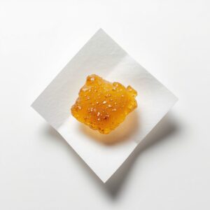 An extreme macro close-up of a small portion of Cannatrek T50 Topaz Bubble Hash concentrate resting on a clean white pharmaceutical weigh paper, soft diffused overhead studio lighting, sterile white laboratory-style background, camera positioned directly overhead at a very slight angle, ultra-high detail rendering capturing the golden-amber colour, surface texture, and trichome-rich consistency of the hash material, clean scientific product documentation photography mood with no lifestyle elements
